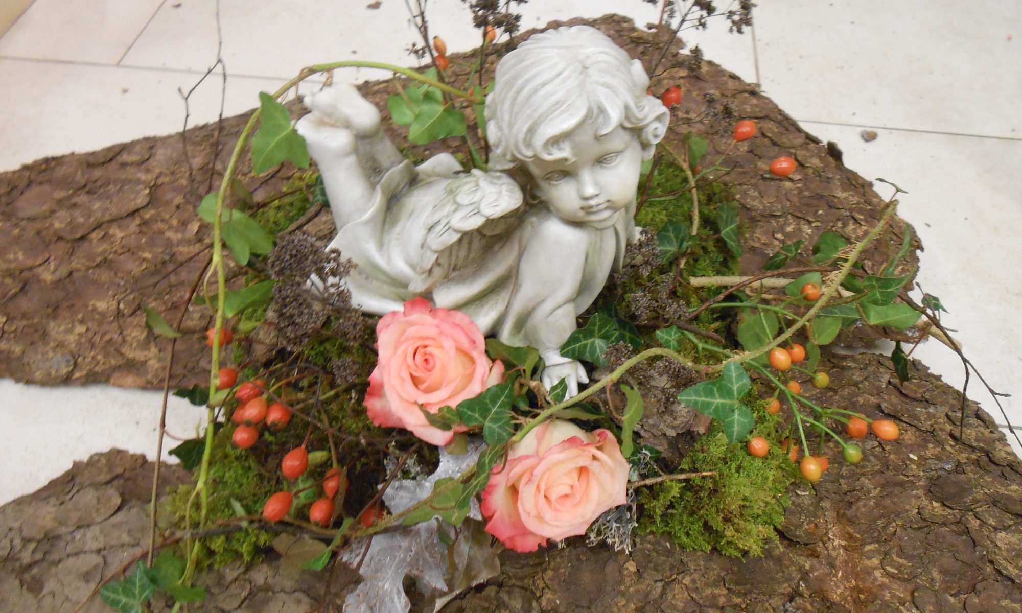 Ein Gesteck auf einer Baumrind bestehend aus zwei rosa Rosen, Hagebutten, Moos, getrockneten Ästen und Efeuranken. In der Mitte vom Gesteck befindet sich ein am Bauch liegender Engel .